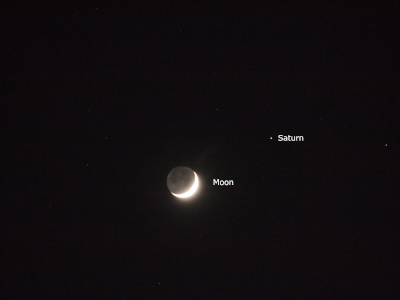 [The sky is black behind the moon and Saturn. Saturn is at two o'clock relative to the moon. Although the white part of the moon is a quarter sliver in the lower right section of the sphere and is very bright, the entire sphere is visible. The non-bright portion of the moon is shades of grey against the black sky and the shades of grey correspond to the valleys of the moon. The name of Saturn and the moon are white text to the right of the orb.]